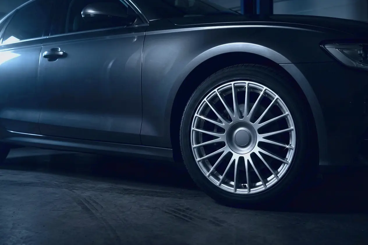 A dark car's front wheel arch and its multi-spoke alloy wheel, lit by cool light in a dark bay at night