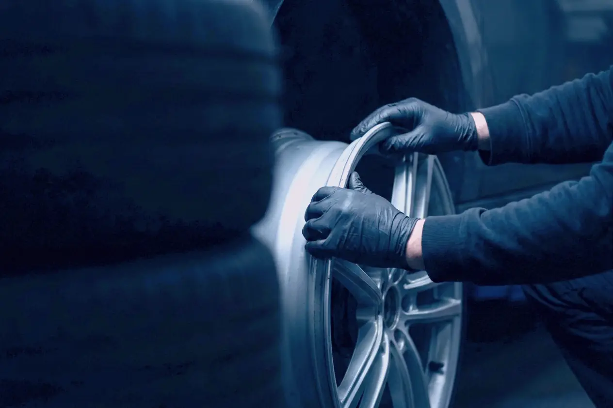 Gloved hands holding a bare alloy rim beside a car in a dark bay