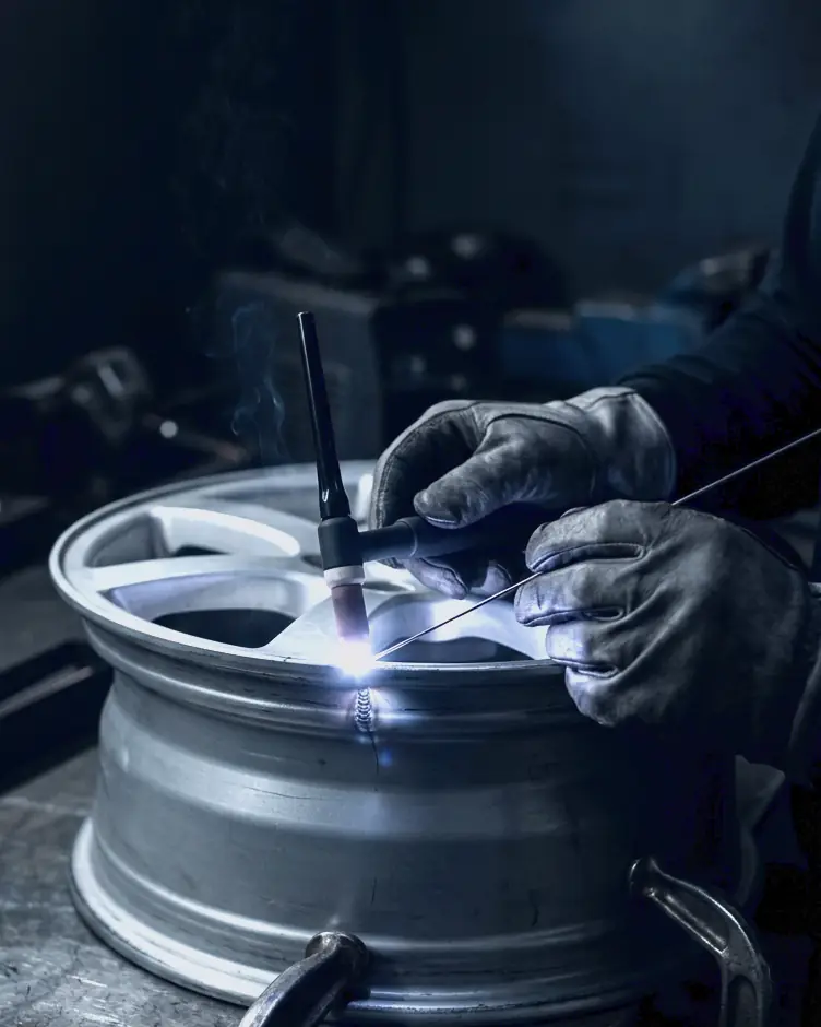 Gloved hands TIG welding a crack on the lip of a bare alloy rim, the arc glowing blue-white